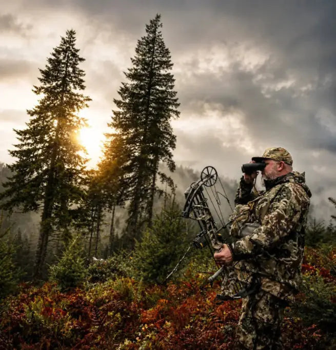 Bowhunter looking through binoculars at sunrise between tall pine trees, silhouetted against a cloudy sky